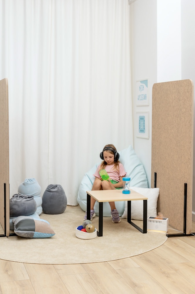 Dziewczynka siedzi w spokojnym kąciku sensorycznym, na pufie, z słuchawkami wygłuszającymi i zabawką sensoryczną w rękach.