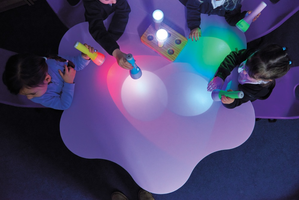 Dzieci bawiące się kolorowymi latarkami przy świetlnym stoliku, tworząc barwne refleksy.